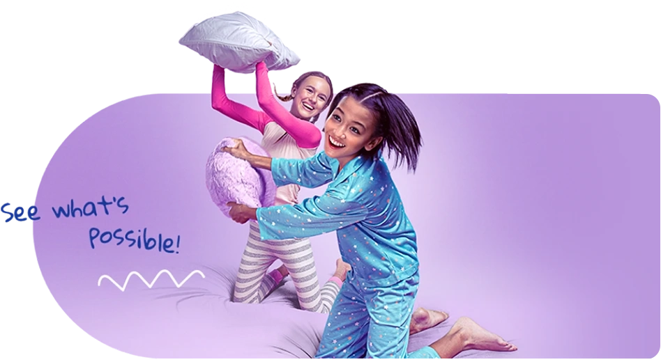 Two girls in pajamas, smiling and holding pillows during a pillow fight. They are next to the words, &quot;See what's possible!&quot;