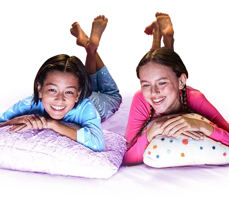 Two girls in pajamas, smiling and resting their arms on their pillows with their feet in the air.