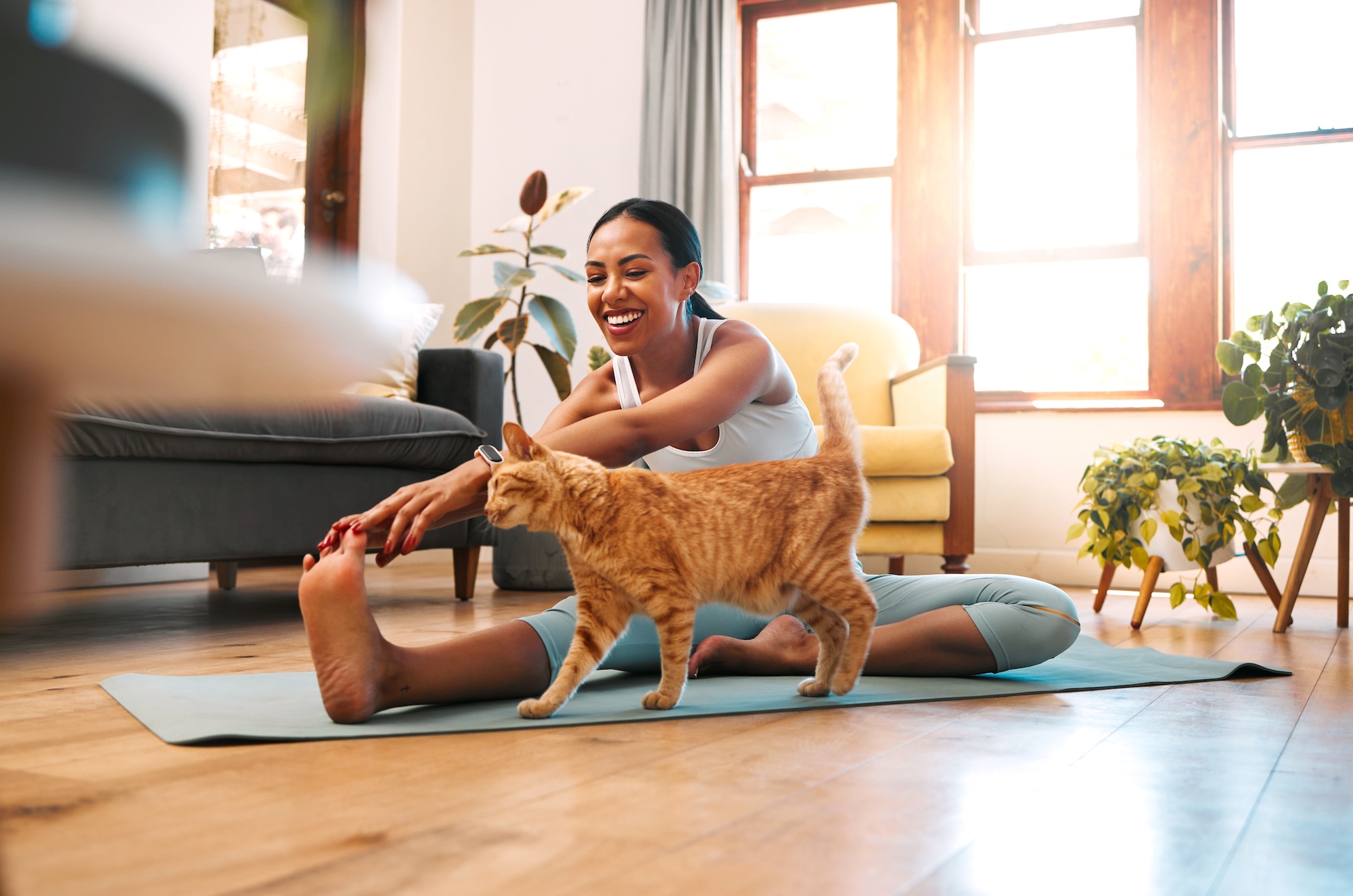 A woman stretching on a yoga mat and smiling as her cat walks by