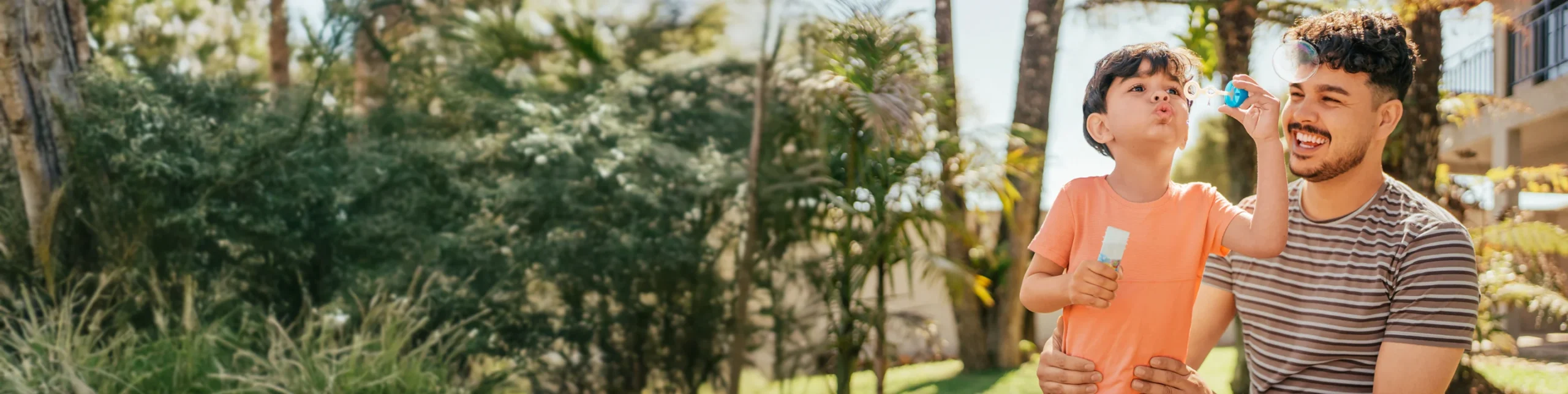 A father holding his hands around his young son's waist as his son blows bubbles outdoors