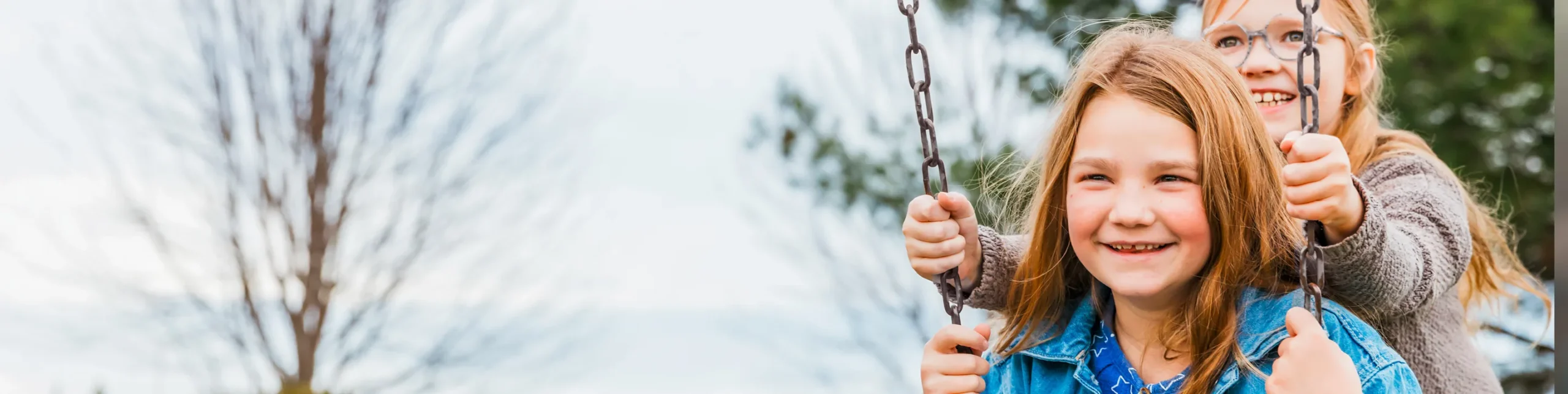 A young girl smiling while being pushed on a swing by her friend outdoors