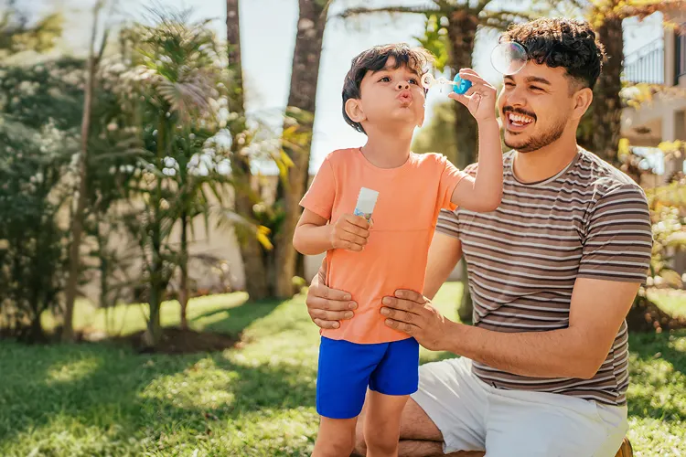 A father holding his hands around his young son's waist as his son blows bubbles outdoors